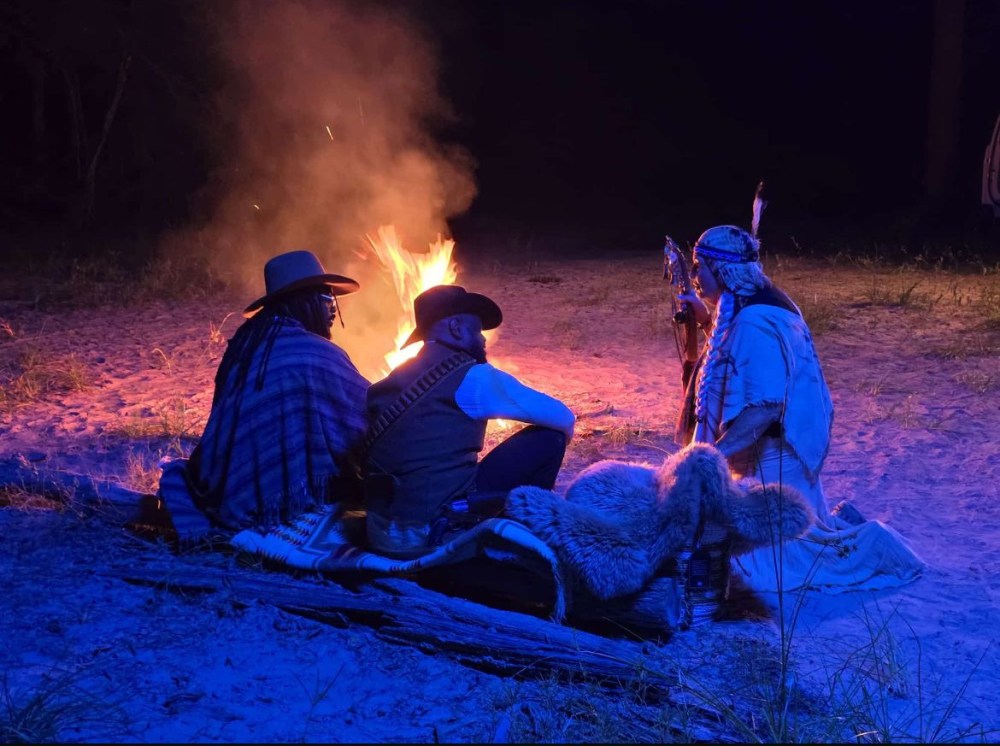 Three people in hats sitting by a campfire at night, surrounded by sand and grass.