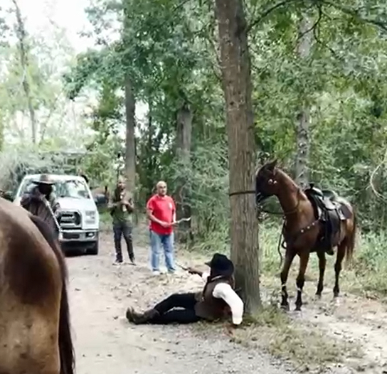 Person in cowboy attire sitting by tree with horse, people and truck in wooded area.