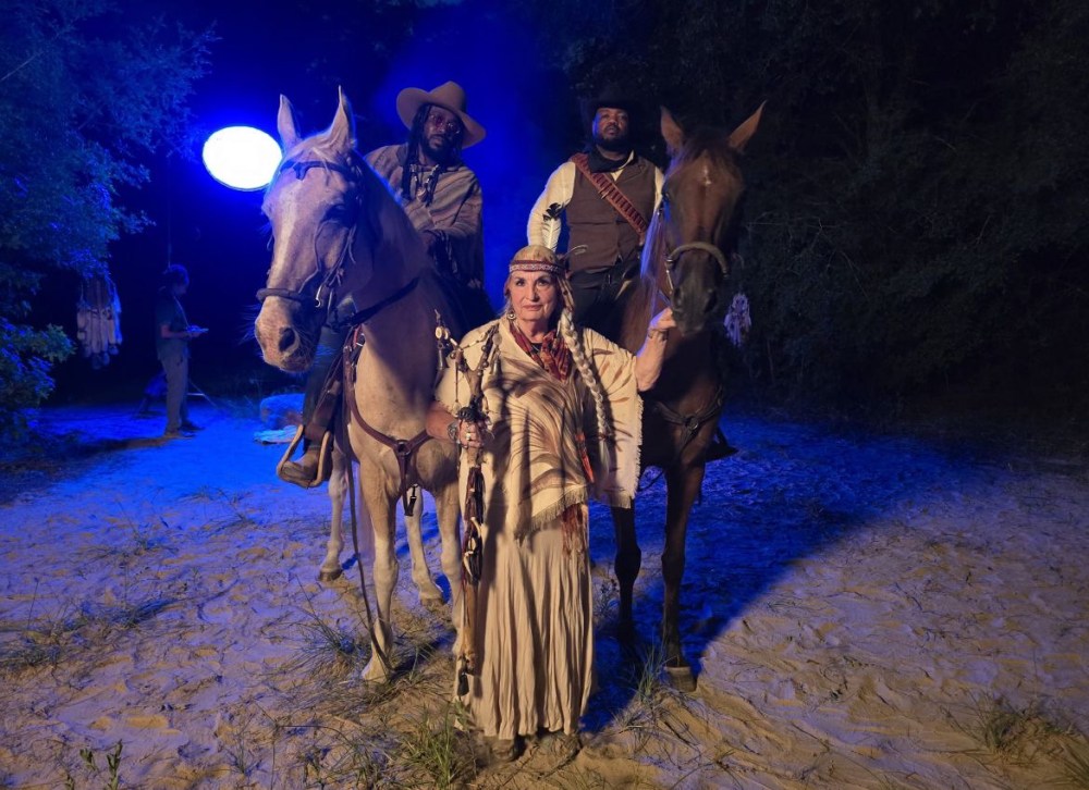 Two men on horses and a woman in costume standing on sandy ground with a blue light.