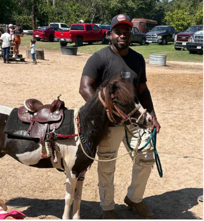Man in black shirt and cap holding a pony on a sunny day with parked trucks in the background.