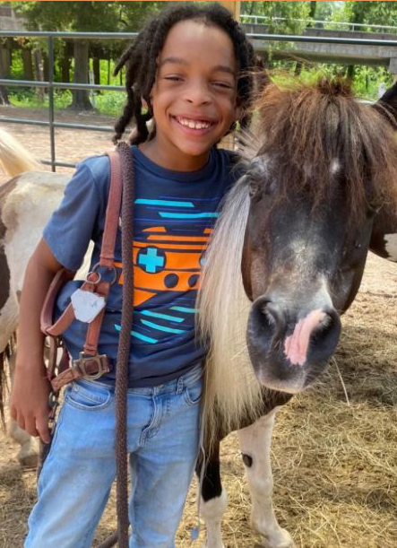 Child smiling next to a pony in a sunny outdoor setting.
