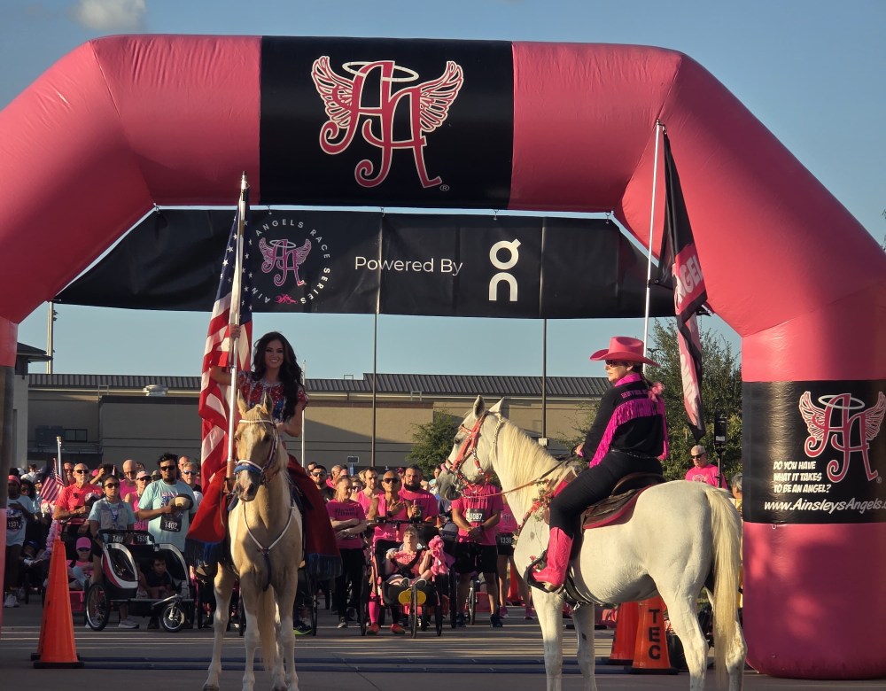 Two horseback riders under pink inflatable arch, one holding an American flag.