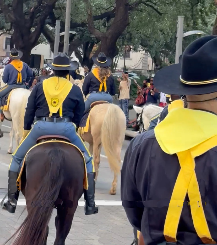People in black uniforms and yellow scarves riding horses, with trees and spectators in the background.