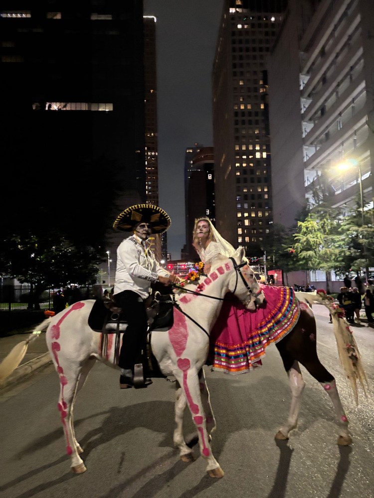Two people in traditional outfits on horses, with painted designs, in a city street at night.