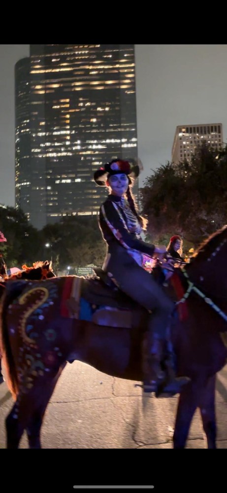 Person in costume with face paint on horse at night, city skyscraper in background.