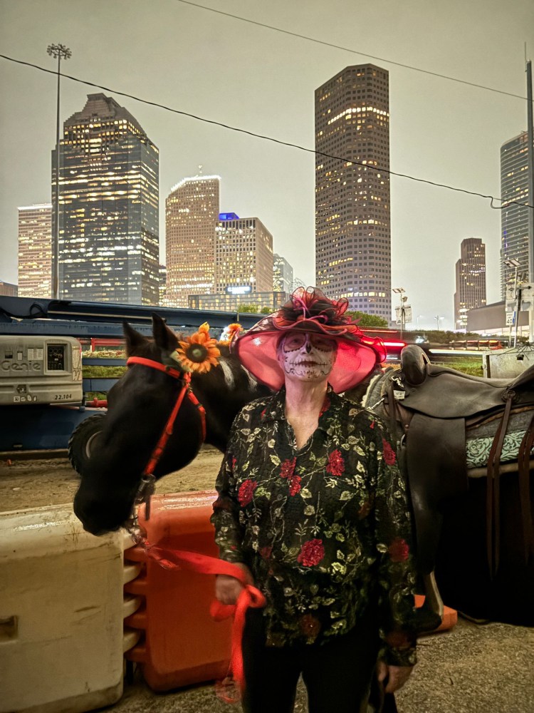 Person in skeleton makeup and floral outfit with horse, city skyline in background at night.