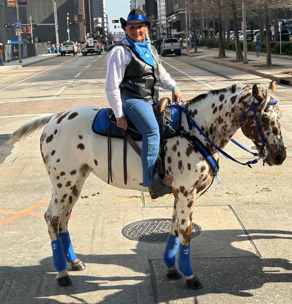 Person on a spotted horse wearing blue accessories and cowboy attire, on a city street.