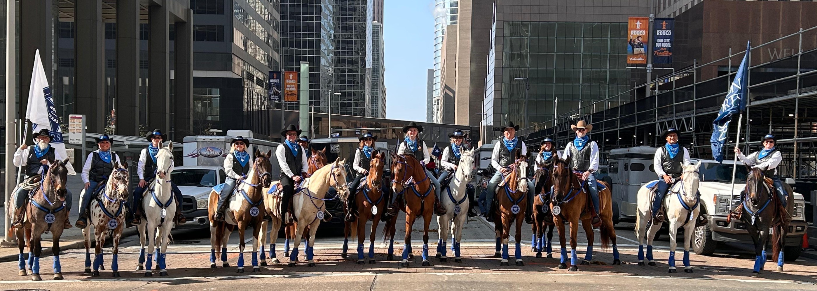 Riders on horses in blue outfits, holding flags on an urban street.