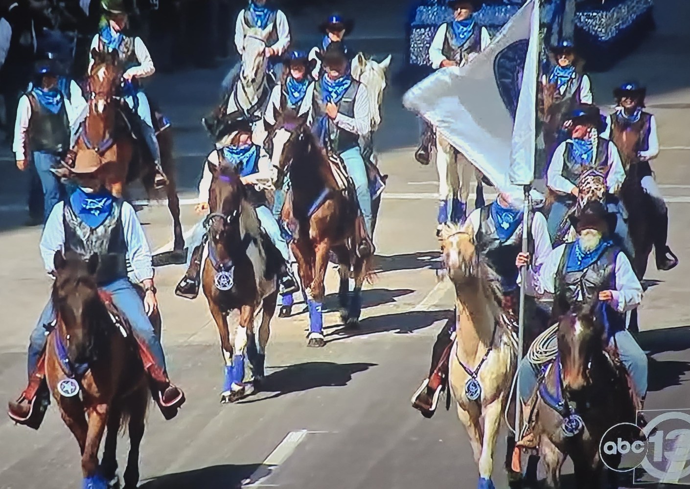Riders in blue bandanas on horses, carrying a flag, participating in a parade.