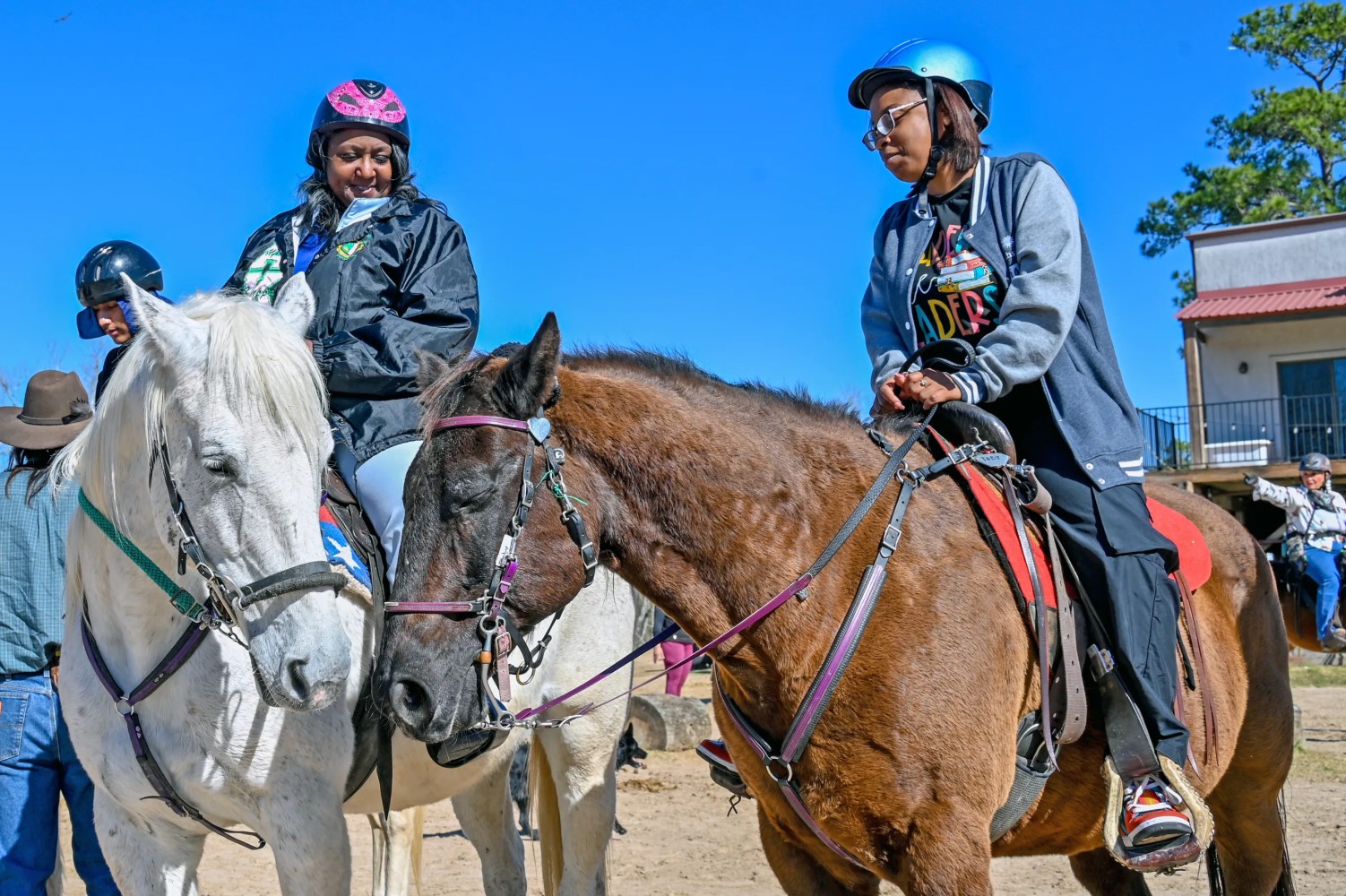 Two people wearing helmets ride horses, talking outdoors on a sunny day.