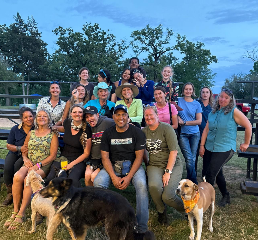 Group of people and two dogs posing outdoors, wooden fence and trees in background.