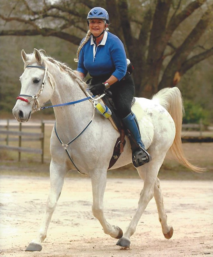 Person in blue riding a white horse outdoors, wearing a helmet.