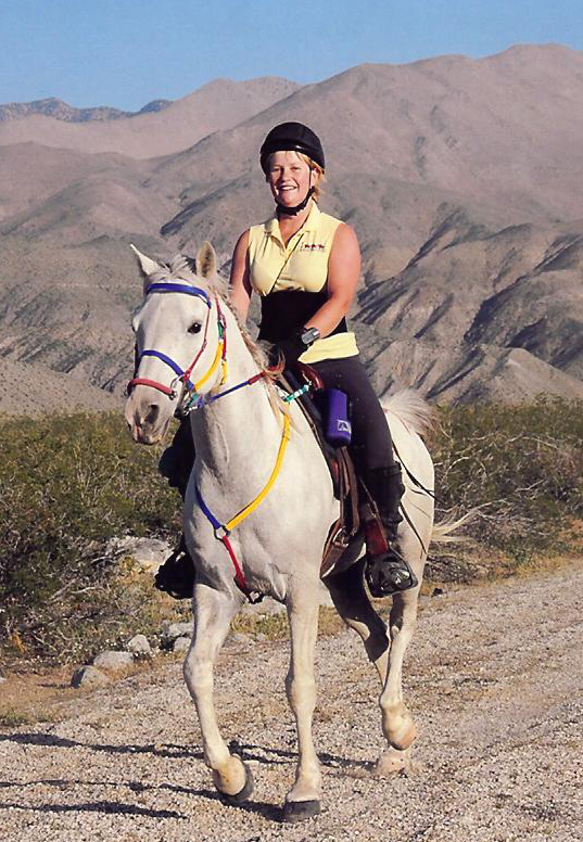 Person riding a white horse on a dirt trail with mountains in the background.