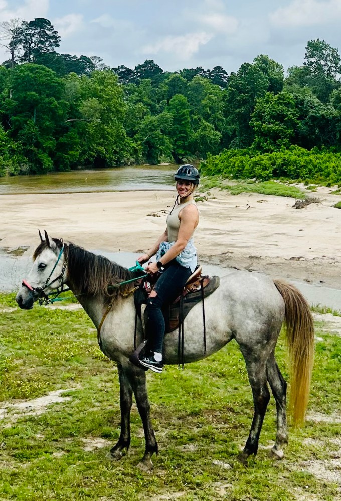 Person on a gray horse by a river, wearing a helmet, sleeveless shirt, and jeans.