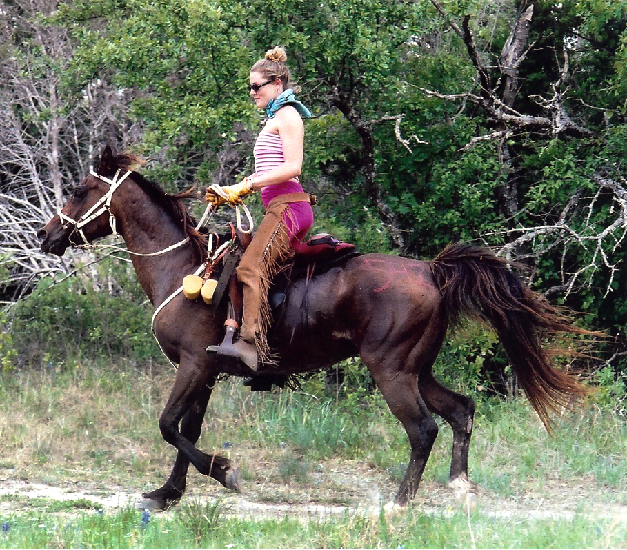 Person riding a horse through a grassy area with trees in the background.