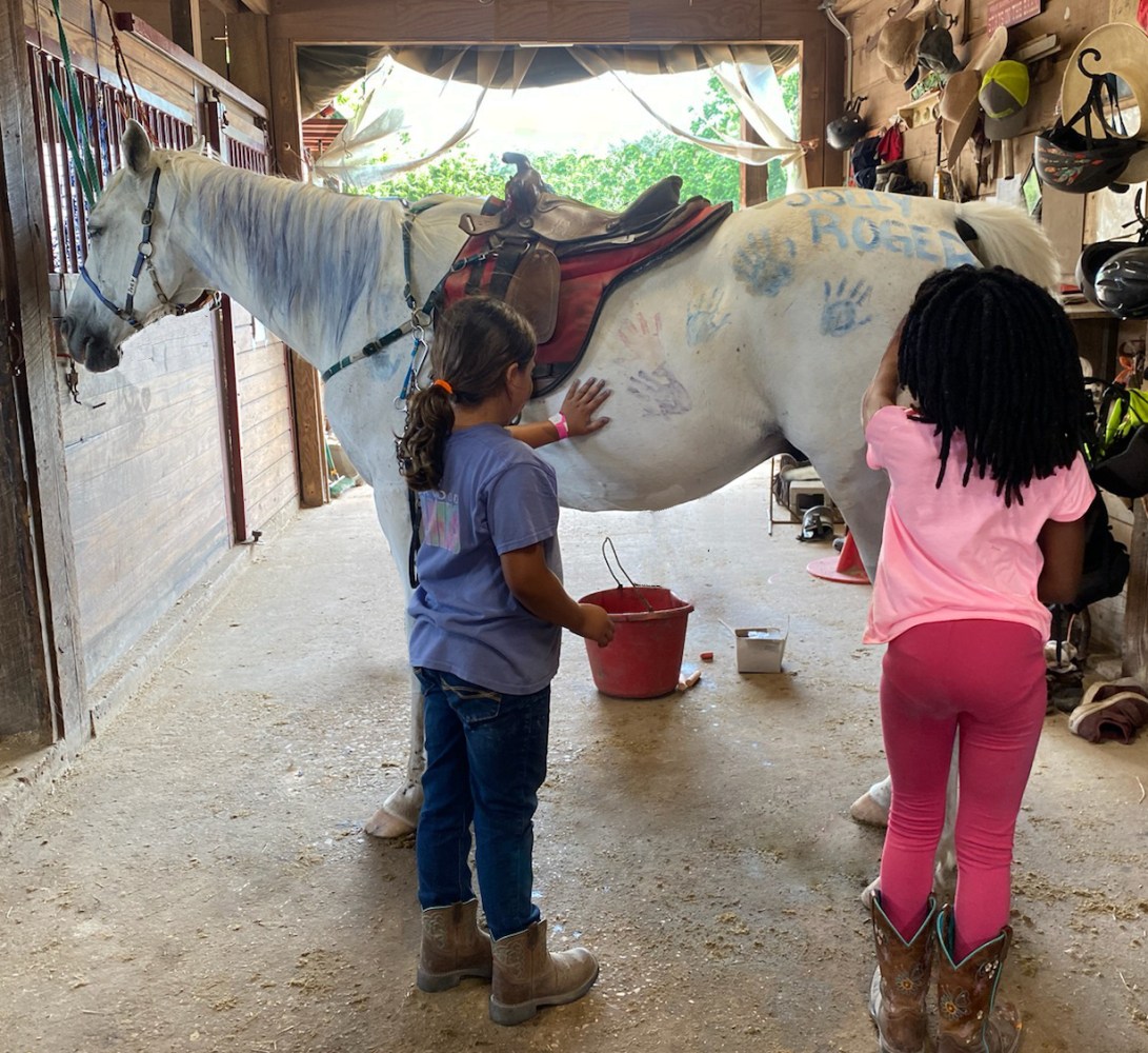 Two children painting a white horse in a stable with colorful handprints.