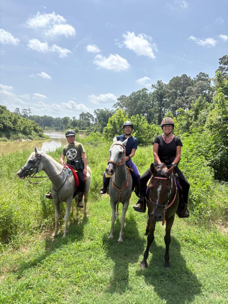 Three people riding horses on a grassy trail by a river.