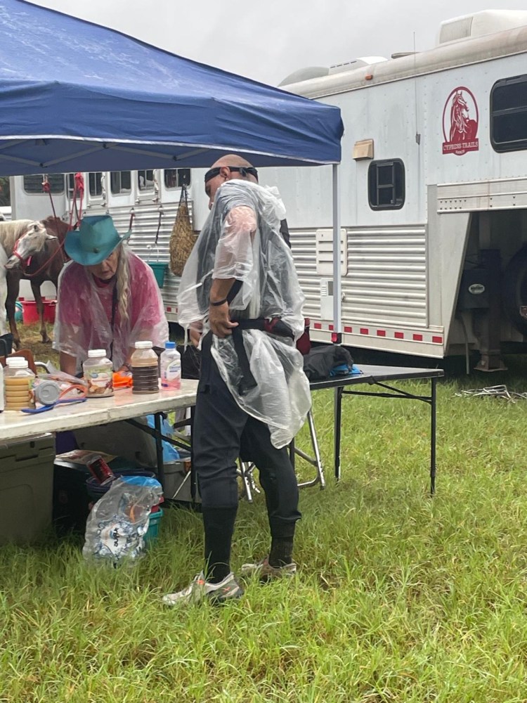 Person in rain poncho under a canopy next to a table with jars, near a horse trailer.